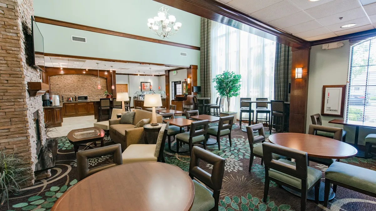 The scene highlights a cozy seating arrangement around a fireplace, complemented by large windows bringing in ample natural light and decorative plants adding a warm touch. The space is designed for relaxation, socializing, or enjoying refreshments in a comfortable environment.