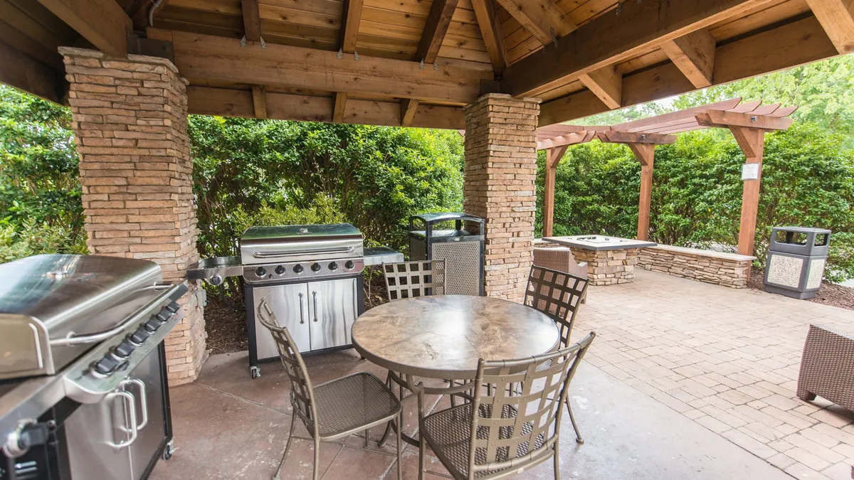 The outdoor barbecue area includes grills, a table set for dining, stone columns, and lush greenery, creating an ideal spot for casual alfresco meals and social gatherings.