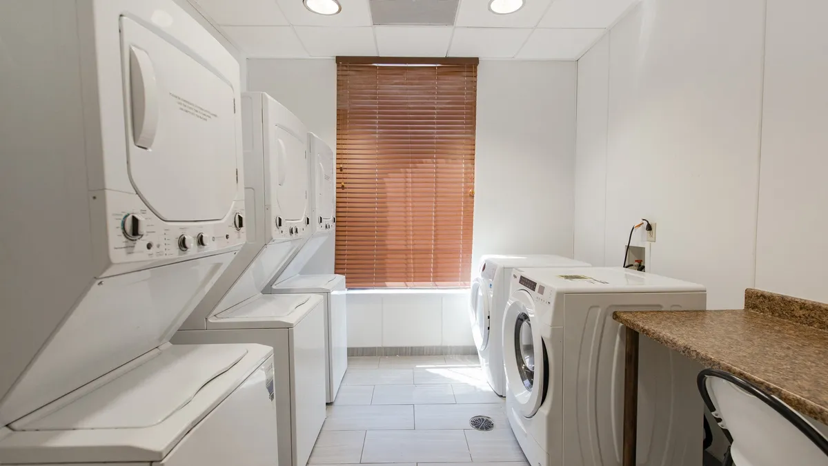 A practical and tidy laundry room featuring multiple stacked washer and dryer units, a countertop surface for folding clothes, and a window with blinds letting in natural light. The room is organized and inviting, emphasizing convenience and cleanliness.