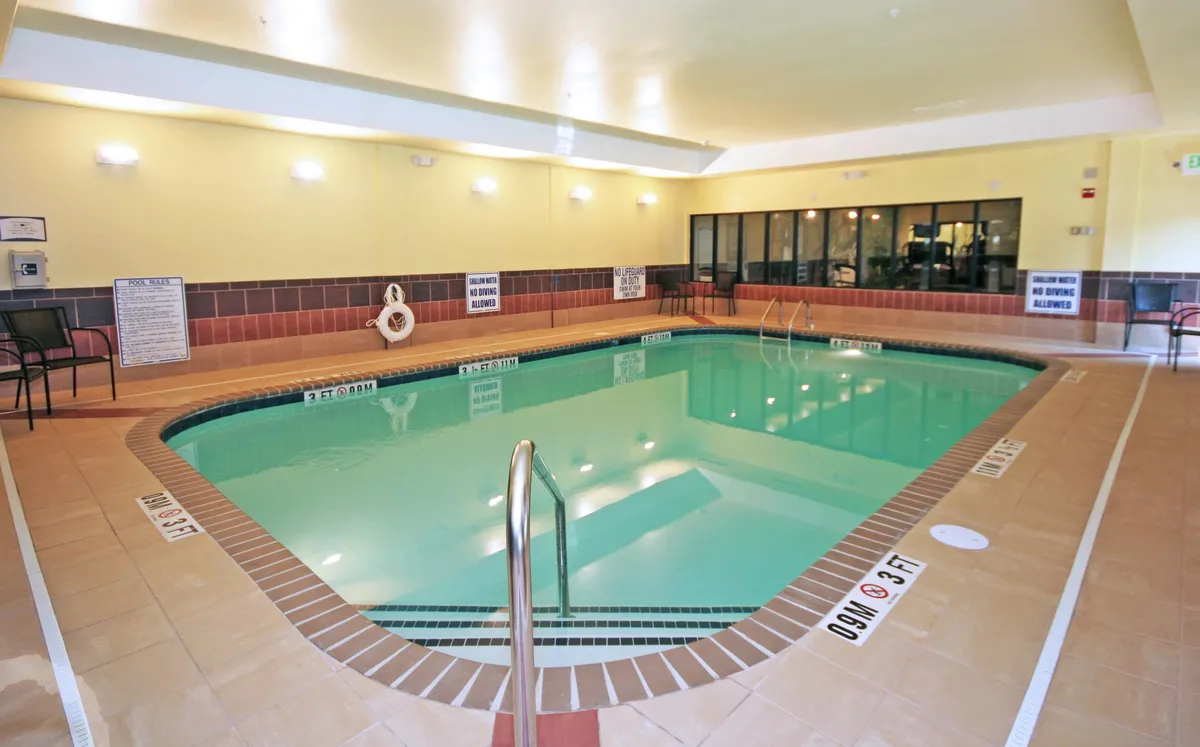 This image captures a spacious indoor pool area, with a ladder leading into the pool, clear safety signage, and surrounding chairs for relaxation. Large windows flood the space with natural light, highlighting the calming blue water. The scene includes areas for swimming, relaxing, and socializing, evoking both activity and leisure.