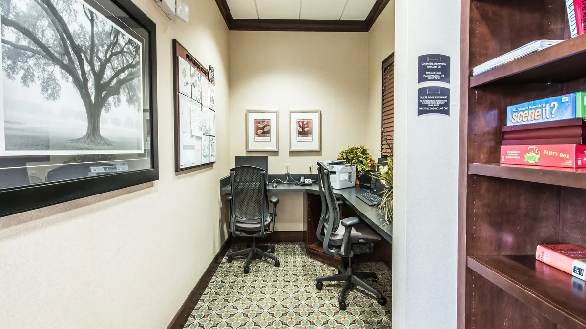 The photo features a modern business center equipped with desks, computers, framed artwork, and a bookshelf. The environment appears quiet and focuses on productivity, with comfortable seating and a clean, organized layout ideal for business travelers or guests needing to work during their stay.