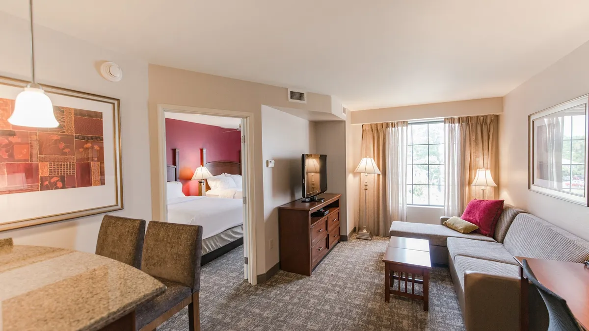 The image shows a cozy and inviting living area featuring a comfortable sofa, a TV stand with a flat-screen TV, a kitchenette, and a window with curtains letting in natural light. The space is arranged for relaxation, socializing, or enjoying a meal, with a warm, home-like ambiance.