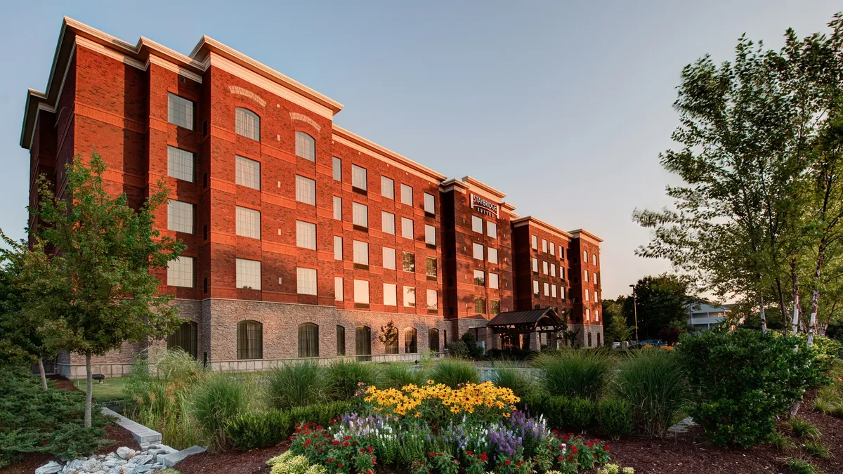 This exterior image features the main entrance canopy of Staybridge Wilmington, surrounded by vibrant flower beds and a classic brick facade. The welcoming outdoor setting invites families to begin their adventures, with lush landscaping and an inviting entrance that hints at a comfortable and lively stay.